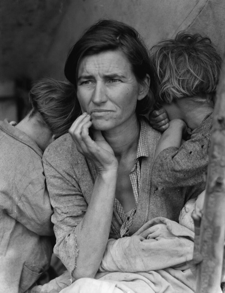 Immagine di una madre seduta attorniata dai bambini con lo sguardo preoccupato e una mano sul viso a lato della bocca. un bambino piccolo nell'altro braccio. Fotografia di Dorothea Lange per la Federal Security Administration degli USA, data 1936.